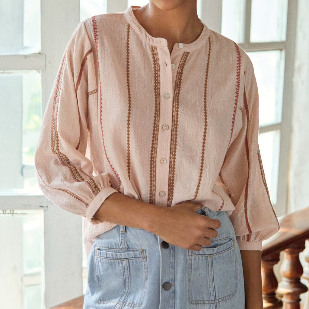 Woman wearing a striped blouse and denim skirt in a bright room with large windows.