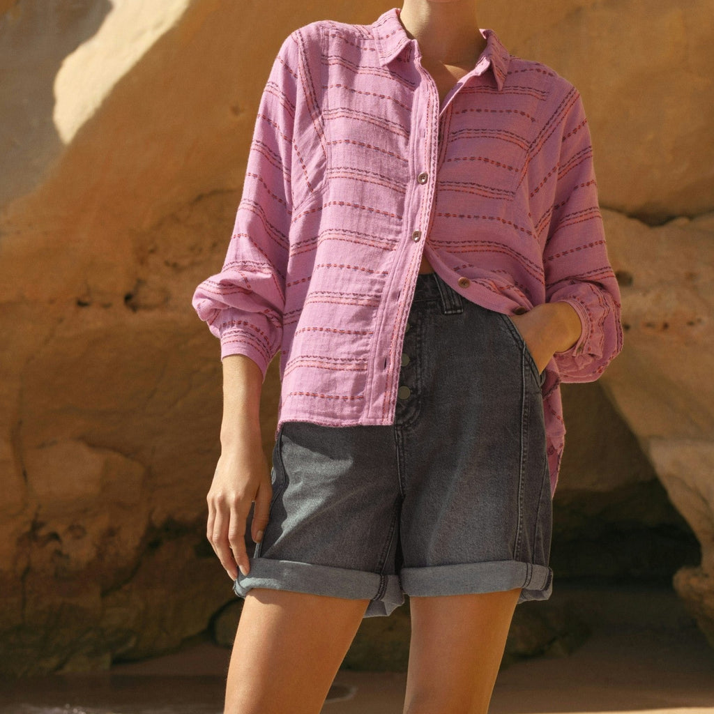 Woman in pink shirt and denim shorts standing on a rocky beach.